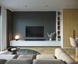 A modern living room with a large flat-screen TV mounted on a dark wall, surrounded by sleek white cabinets. A globe-shaped lamp emits a soft light from a cabinet, and decorative items, including a vase with pampas grass, adorn the space. A neutral-toned L-shaped sofa faces the TV, adjacent to a small round coffee table with books on it. The room has light wooden floors arranged in a herringbone pattern, and a warm wooden paneling contrasts with the contemporary decor. Shelves display books and items, contributing to an organized yet cozy atmosphere.