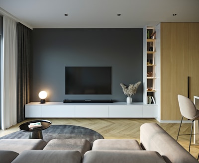 A modern living room with a large flat-screen TV mounted on a dark wall, surrounded by sleek white cabinets. A globe-shaped lamp emits a soft light from a cabinet, and decorative items, including a vase with pampas grass, adorn the space. A neutral-toned L-shaped sofa faces the TV, adjacent to a small round coffee table with books on it. The room has light wooden floors arranged in a herringbone pattern, and a warm wooden paneling contrasts with the contemporary decor. Shelves display books and items, contributing to an organized yet cozy atmosphere.