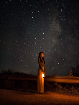 A person stands by a dark road holding a glowing lantern under a sky filled with stars and the Milky Way. The scene is dimly lit, emphasizing the starry night sky and the warm glow of the lantern.