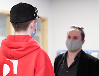 Two individuals wearing face masks are in conversation. One person is wearing a bright red hoodie and a black cap, while the other is dressed in darker clothing with sunglasses perched on their head. The background is blurred, giving focus to the people in the foreground.