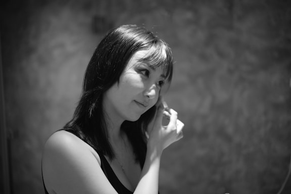 A person with medium-length hair is applying makeup or using a cosmetic tool on their face. The lighting creates soft shadows, and the background appears to be textured.