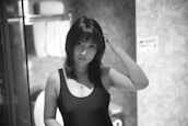 A black and white photo of a woman with shoulder-length dark hair, wearing a sleeveless top. She is touching her hair with one hand and standing in a bathroom with towels visible in the background.