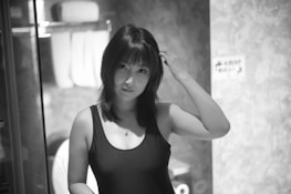 A black and white photo of a woman with shoulder-length dark hair, wearing a sleeveless top. She is touching her hair with one hand and standing in a bathroom with towels visible in the background.