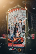A vibrant and ornate depiction of Hindu deities, including a powerful goddess standing atop a lion. The figures are adorned with intricate jewelry and garments, set against a richly decorated backdrop. The atmosphere is ceremonial, enhanced by warm light and floating particles, evoking a sense of reverence and festivity.