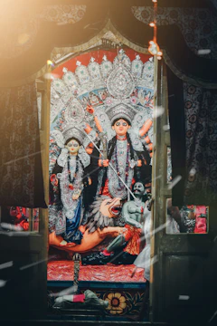 A vibrant and ornate depiction of Hindu deities, including a powerful goddess standing atop a lion. The figures are adorned with intricate jewelry and garments, set against a richly decorated backdrop. The atmosphere is ceremonial, enhanced by warm light and floating particles, evoking a sense of reverence and festivity.