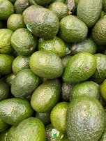 A pile of avocados with smooth and bumpy green skins, some bearing stickers indicating their origin or type. The avocados are tightly packed together, displaying varying shades of green.