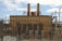 A large industrial power plant with two prominent smokestacks rises in the background, adorned with the word 'POWER' in bold red letters. The foreground is dominated by a complex maze of electrical grid structures, wires, and transformers. Chain-link fences and caution signs suggest restricted access to the facility. The sky above is partly cloudy, offering a contrast to the industrial scene below.