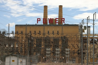 A large industrial power plant with two prominent smokestacks rises in the background, adorned with the word 'POWER' in bold red letters. The foreground is dominated by a complex maze of electrical grid structures, wires, and transformers. Chain-link fences and caution signs suggest restricted access to the facility. The sky above is partly cloudy, offering a contrast to the industrial scene below.