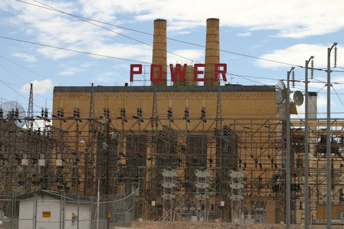 A large industrial power plant with two prominent smokestacks rises in the background, adorned with the word 'POWER' in bold red letters. The foreground is dominated by a complex maze of electrical grid structures, wires, and transformers. Chain-link fences and caution signs suggest restricted access to the facility. The sky above is partly cloudy, offering a contrast to the industrial scene below.