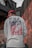 A person wearing a white shirt with a graphic design featuring skulls and the text 'OFF THE WALL' is standing with their back to the camera. They are also wearing a red cap. The background consists of urban elements, including a concrete wall and some metal structures.