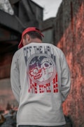 A person wearing a white shirt with a graphic design featuring skulls and the text 'OFF THE WALL' is standing with their back to the camera. They are also wearing a red cap. The background consists of urban elements, including a concrete wall and some metal structures.