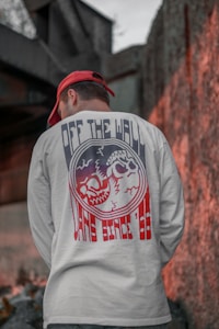 A person wearing a white shirt with a graphic design featuring skulls and the text 'OFF THE WALL' is standing with their back to the camera. They are also wearing a red cap. The background consists of urban elements, including a concrete wall and some metal structures.