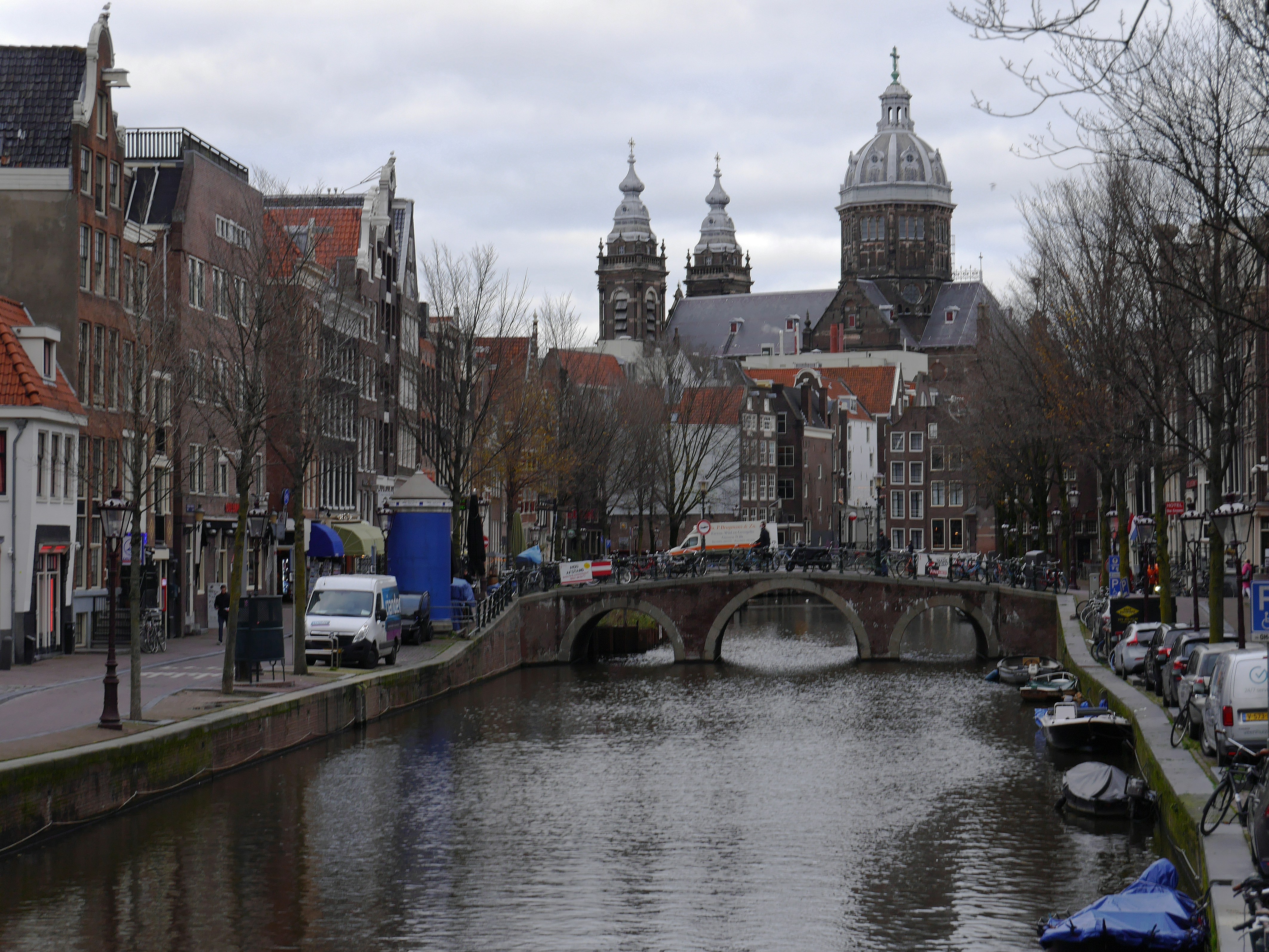 Amsterdam canal city - street photography from Holland. A street view over the old canal with in the background the towers of the old church Sint Nicolas, located at the border of the red light district - downtown. The canal water is reflecting the gray sky and the house facades in winter colors. Free pic photo of churches in Amsterdam city, pictures of The Netherlands by Dutch photographer Fons Heijnsbroek. dutch: straatfotografie Amsterdam, Nederland, hoge resolutie en rechtenvrij