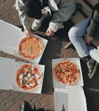three people sitting on a bench with three pizzas in boxes