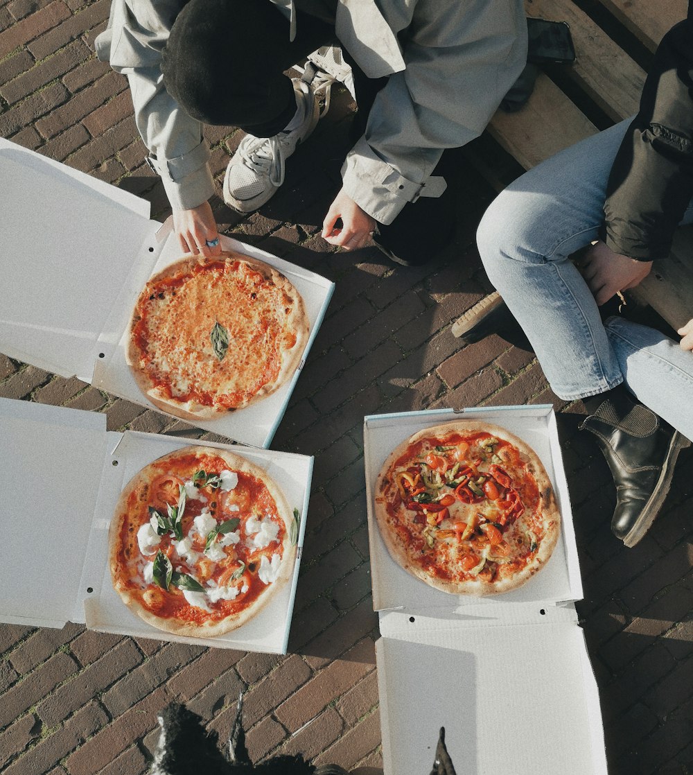 three people sitting on a bench with three pizzas in boxes