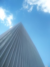 A tall, sleek building with a modern design and vertical lines reaches up towards a bright blue sky with scattered clouds.