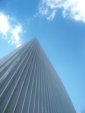 A tall, sleek building with a modern design and vertical lines reaches up towards a bright blue sky with scattered clouds.