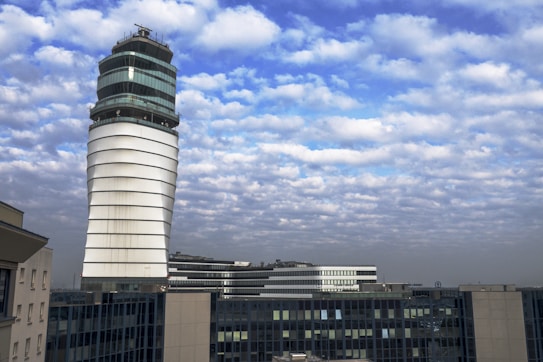 A modern airport control tower with a unique, spiral design rises prominently against a partly cloudy sky. Surrounding the tower are other airport buildings with large glass windows, reflecting the daylight.