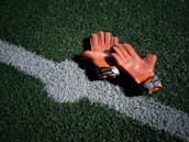 A pair of worn orange goalkeeper gloves resting on a green artificial turf field, positioned beside a white line.
