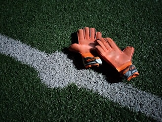 A pair of worn orange goalkeeper gloves resting on a green artificial turf field, positioned beside a white line.