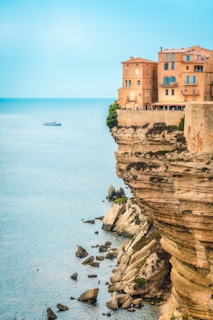 A picturesque seaside cliff with tall, rustic buildings perched at the edge, overlooking the vast blue sea. A small boat sails calmly on the water near the horizon, while the rocky cliffside descends steeply to the ocean below. The scene is serene, with minimal foliage, emphasizing the architectural and natural beauty.