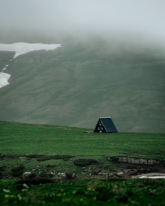 A small, A-frame cabin is situated on a vast, lush green meadow. Misty hills rise in the background, partially covered by patches of lingering snow. The atmosphere is calm and serene, emphasizing solitude and nature's tranquility.