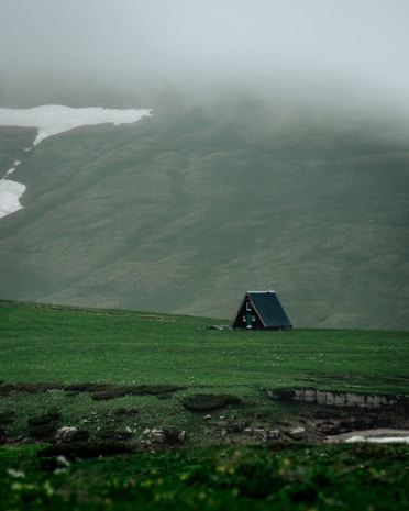 A small, A-frame cabin is situated on a vast, lush green meadow. Misty hills rise in the background, partially covered by patches of lingering snow. The atmosphere is calm and serene, emphasizing solitude and nature's tranquility.