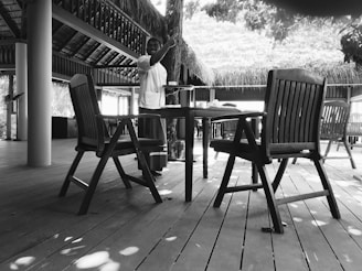 A person wearing a uniform is standing next to a wooden table and chairs on a deck with a thatched roof and natural light filtering through leaves. The setting appears to be outdoors or a semi-open space with a tropical or rustic vibe.