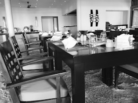 A dining area with neatly arranged tables and chairs. The tables are set with tableware, including glasses and napkins. The space has a modern aesthetic with clean lines and decorative elements on the walls. Overhead lighting is visible, creating a bright and welcoming environment.