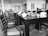 A dining area with neatly arranged tables and chairs. The tables are set with tableware, including glasses and napkins. The space has a modern aesthetic with clean lines and decorative elements on the walls. Overhead lighting is visible, creating a bright and welcoming environment.