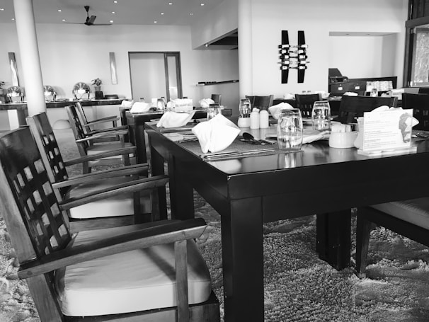 A dining area with neatly arranged tables and chairs. The tables are set with tableware, including glasses and napkins. The space has a modern aesthetic with clean lines and decorative elements on the walls. Overhead lighting is visible, creating a bright and welcoming environment.