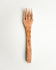 A wooden fork with a unique, textured design featuring elongated, pointed tines. The fork displays natural wood grain patterns that provide a rustic and artistic appearance. It is set against a pale, neutral background.