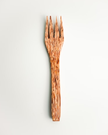 A wooden fork with a unique, textured design featuring elongated, pointed tines. The fork displays natural wood grain patterns that provide a rustic and artistic appearance. It is set against a pale, neutral background.