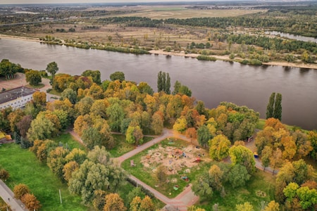 An aerial view of a large park with numerous trees, some of which exhibit autumn colors. The park is bordered by a wide river on the right side, with areas of open land and more trees stretching into the distance. Within the park, there is a playground with various play equipment. Surrounding the park are pathways and a building can be seen on the left side among the trees.
