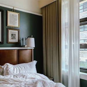 A cozy bedroom with a white unmade bed and a brown leather headboard. There are framed artworks on a green wall, including a playbill. A white table lamp is attached to the wall. A window with sheer white and beige curtains lets in natural light, revealing an exterior view of a brick building.