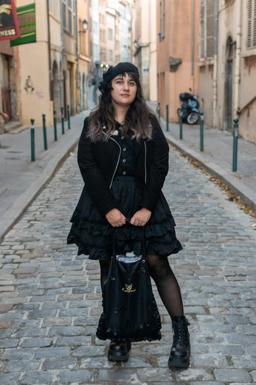 a woman in a black dress and hat is standing on a cobblestone street