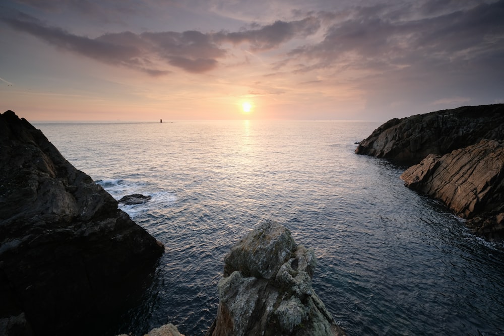 the sun is setting over the ocean with rocks in the foreground