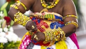 A person adorned in vibrant, traditional African attire featuring a variety of intricate patterns and bright colors. The individual is wearing an abundance of gold jewelry, including bracelets, rings, and necklaces with rich details. Their outfit includes a mix of yellow, blue, red, and green in traditional designs. The background shows a blurred floral arrangement.