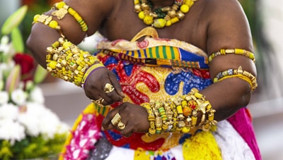 A person adorned in vibrant, traditional African attire featuring a variety of intricate patterns and bright colors. The individual is wearing an abundance of gold jewelry, including bracelets, rings, and necklaces with rich details. Their outfit includes a mix of yellow, blue, red, and green in traditional designs. The background shows a blurred floral arrangement.