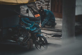 A person is working on the ground next to a damaged motorized rickshaw with a 'Fuera de Servicio' sign. The scene appears to be taking place in an outdoor setting with a brick wall in the background.