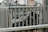 A stack of firewood is neatly arranged behind a wooden railing on a porch. The setting includes a wooden patio chair and a couple of tables, suggesting a cozy, outdoor space. The railing and furniture are weathered, giving a rustic feel to the area.