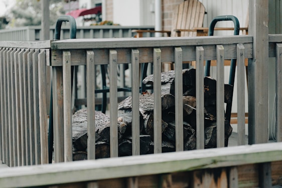 A stack of firewood is neatly arranged behind a wooden railing on a porch. The setting includes a wooden patio chair and a couple of tables, suggesting a cozy, outdoor space. The railing and furniture are weathered, giving a rustic feel to the area.