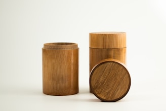 Three cylindrical bamboo containers are arranged on a plain, light background. One container is open, revealing its hollow interior, while the other is closed with a lid. A separate, round lid is placed on its side in front of the containers, showcasing the wood grain pattern.