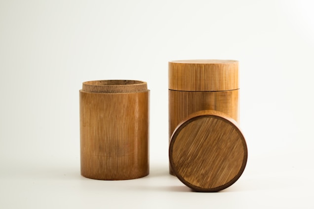 Three cylindrical bamboo containers are arranged on a plain, light background. One container is open, revealing its hollow interior, while the other is closed with a lid. A separate, round lid is placed on its side in front of the containers, showcasing the wood grain pattern.