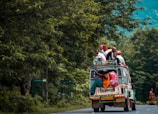 A vehicle is traveling down a road with several people sitting on the roof and hanging onto the back. The vehicle is surrounded by dense, lush greenery. The attire of the people is colorful, and they appear to be crowded together on the vehicle.