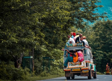 A vehicle is traveling down a road with several people sitting on the roof and hanging onto the back. The vehicle is surrounded by dense, lush greenery. The attire of the people is colorful, and they appear to be crowded together on the vehicle.