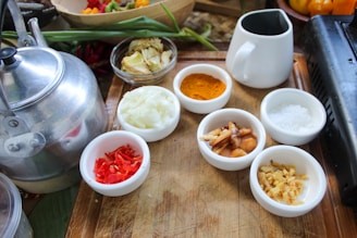 A wooden tray holds various small white bowls filled with ingredients including chopped onions, red chili peppers, turmeric powder, salt, and sliced ginger. A silver kettle is placed on the left side along with a white jug. Green onions and other vegetables are visible in the background.