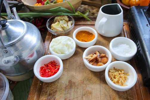 A wooden tray holds various small white bowls filled with ingredients including chopped onions, red chili peppers, turmeric powder, salt, and sliced ginger. A silver kettle is placed on the left side along with a white jug. Green onions and other vegetables are visible in the background.