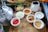 A wooden tray holds various small white bowls filled with ingredients including chopped onions, red chili peppers, turmeric powder, salt, and sliced ginger. A silver kettle is placed on the left side along with a white jug. Green onions and other vegetables are visible in the background.