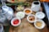 A wooden tray holds various small white bowls filled with ingredients including chopped onions, red chili peppers, turmeric powder, salt, and sliced ginger. A silver kettle is placed on the left side along with a white jug. Green onions and other vegetables are visible in the background.